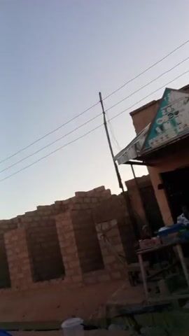 The video depicts a street scene in a rural area, likely during sunset or sunrise. The camera moves, capturing views of buildings, some of which appear to be shops or small businesses with people sitting outside.  A woman in a light green headscarf and dress walks by on the street. The road is unpaved, and there are several motorbikes parked nearby. The sun is visible in the background.