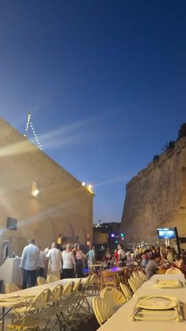 This video shows an outdoor gathering at dusk with a clear, dark blue sky. In the foreground, several long white tables are set with white chairs and place settings. Beyond the tables, a crowd of people are milling around and seated. To the left, a building with arched openings is illuminated by warm lighting, with a string of lights overhead. To the right, a large, stone fortification wall dominates the background. Some people are dancing in front of a stage area, while others are seated at tables, eating and socializing. The atmosphere appears festive, with a sense of community and celebration. The scene is set in an urban or historical environment.
