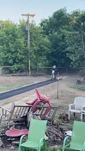 A motocross rider is shown performing a jump. The video begins with the rider approaching a ramp on a dirt track. They ascend the ramp and launch into the air, performing a trick while airborne. The rider then descends towards an inflatable landing ramp. The environment appears to be a backyard or rural setting, with trees and a fence visible in the background.