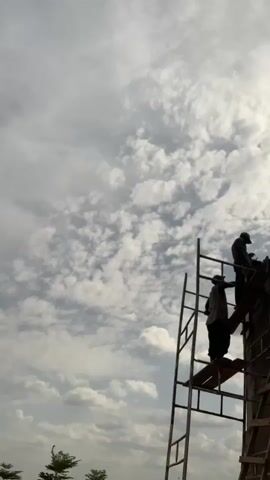 Two construction workers are on scaffolding, working on a brick building under a cloudy sky. One worker, wearing a green helmet, is at the top of the structure, while the other, in a hat, stands lower on the scaffolding. They are both silhouetted against the sky. Some greenery is visible at the bottom left.