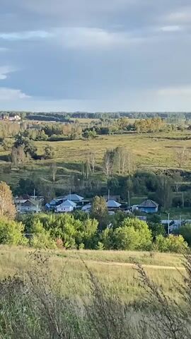The video shows a scenic view of a rural landscape. In the foreground, tall grasses sway in the breeze. Beyond the grasses, a village is nestled in a valley. Houses with colorful roofs are visible amongst lush green trees. In the distance, rolling hills stretch towards the horizon, dotted with more trees and buildings. The sky above is partly cloudy, with patches of blue sky peeking through. The overall impression is one of peace and tranquility in a natural setting.