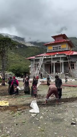 A group of people, mostly women wearing traditional clothing, are working in an outdoor area. Some are digging or moving materials, while others appear to be observing or coordinating. A building with a red roof and scaffolding is visible in the background, suggesting construction or renovation is underway. The environment is mountainous and green, with some clouds in the sky. There are also some smaller structures and a prayer flag in the vicinity. The overall scene depicts a community effort in a rural, possibly remote, setting.