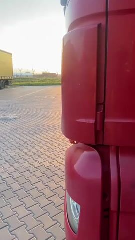 The video is a close-up, low-angle shot of the side of a red semi-truck cab from a driver's perspective. The camera is positioned close to the truck, showing the side panel and the front fender with a portion of the headlight. The truck is parked on a paved lot made of interlocking concrete pavers. To the left of the truck, in the background, is a yellow semi-trailer with the text "OMEGA Pizza LOGISTICS" visible. The sun is setting on the horizon, casting a warm orange glow and lens flares across the scene. The sky is a light blue with wispy clouds. In the distance, there are faint outlines of structures and possibly airport facilities. The video shows no people or any action.