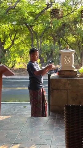 A man wearing a dark t-shirt and a patterned sarong walks in front of a restaurant, carrying a tray of items. He stops at an outdoor altar with a white stone lantern and appears to place offerings on it. The scene is set outdoors with lush green trees and dappled sunlight.