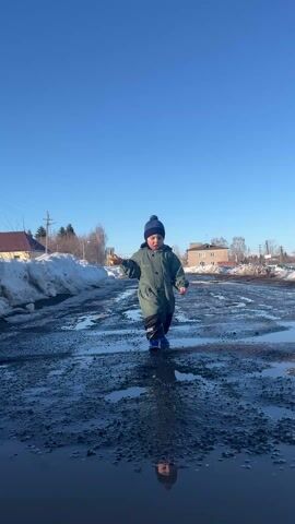 A young child in a green winter jumpsuit, blue boots, and a blue knit hat walks through a muddy, pothole-filled road on a sunny day. The child is holding a small, round, brown object in their left hand. The road is wet, with many puddles reflecting the child and the clear blue sky.  Patches of melting snow are visible along the sides of the road, and some buildings can be seen in the background. The child walks forward with a slightly unsteady gait, navigating the puddles. The video is shot from a low angle, emphasizing the puddles and the child's interaction with them.