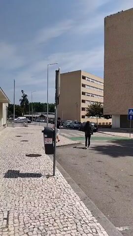 The video begins with a view of a paved area with a cobblestone sidewalk on the left and a street on the right. A tall, dark trash receptacle is visible on the sidewalk. A metal pole with a weathered sign is also on the sidewalk. Two people are walking across a crosswalk. The person closer to the camera is wearing dark clothing and a backpack, and is walking away from the camera. The person further from the camera is wearing a light-colored shirt and shorts and is also walking away from the camera. Behind the crosswalk, there are several cars parked. In the background, there are two large, multi-story buildings. The building on the right is a tall, rectangular structure made of brick. The building on the left is lighter in color and has many windows. The sky is blue with some clouds. The scene appears to be an urban or suburban environment on a sunny day. The video then pans slightly to the right and upward, focusing more on the brick building and the sky. The camera continues to move, capturing more of the buildings and the street. Finally, the camera moves to the left, showing a different angle of the cobblestone sidewalk and more of the surrounding environment, including trees and streetlights. The video concludes with a wider view of the area, showcasing the buildings, cars, and the sky.