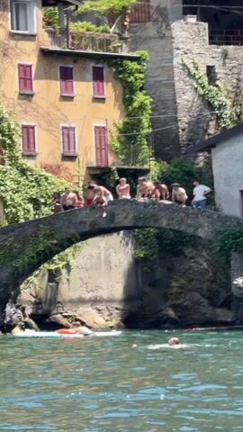 A group of young men are gathered on a stone bridge, some of them preparing to jump into the water below. One person has already jumped and is mid-air. Several other people are in the water, some on paddleboards and others swimming. The bridge spans a body of water next to buildings with vibrant yellow and stone facades, covered in lush green vines. The scene appears to be a warm, sunny day.