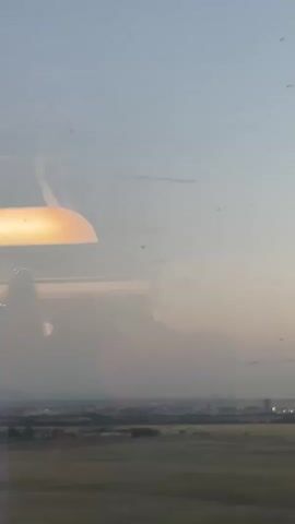 A view from inside a plane shows a hazy landscape through the window.  The sky is light blue with a slight gradient towards the horizon. In the distance, there's a stretch of land with a mix of grassy areas and some buildings or structures clustered together. A light source, possibly an interior light, is reflected in the window. The window itself has some minor smudges or dirt particles visible.