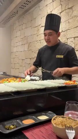 The video shows a male chef in a black chef's coat and a tall black chef's hat preparing food on a teppanyaki grill. He is smiling and using a spatula and tongs to move food, which appears to be chicken and vegetables, onto white rectangular plates. Steam rises from the grill. In the foreground, there are three small black dishes containing sauces or condiments, a glass of water, and a bowl of rice. The background features a stone wall and a metal exhaust hood above the grill. The lighting is warm and indoor.