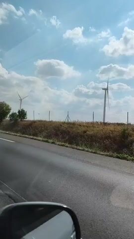 A video shows a roadside view from a car, focusing on wind turbines in the distance. The camera is pointed out the side window, capturing a highway with a white line down the middle. To the right of the road, there's a dry, grassy field with several trees and fence posts. The sky is blue with scattered white clouds. Three wind turbines are visible in the field; one is closer and larger, while two others are further away and smaller. The reflection of the car's side mirror is visible in the bottom corner of the frame. The video appears to be a continuous recording without any significant action or interaction.