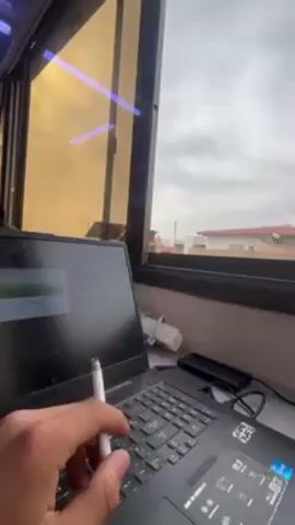The video shows a person sitting next to a window, using a laptop.  The laptop is black and has an ASUS sticker on it. A charging symbol is displayed on the screen. The person is holding a white object, potentially a pen or stylus, near the keyboard. Next to the laptop, there is a white cable organizer, a black hard drive, and a pair of glasses. The window has a black frame with yellow-tinted glass. Outside the window, the view is of a neighborhood with residential buildings and a cloudy sky. Solar panels are visible on the roof of one of the buildings.