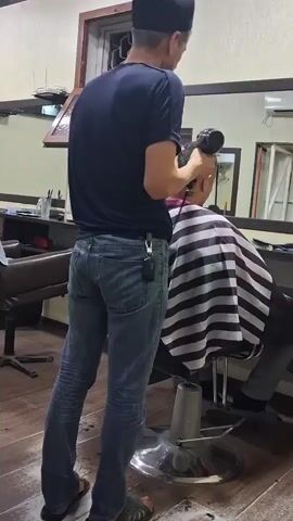 A man in a blue t-shirt and jeans is blow-drying the hair of a person sitting in a barber chair. The person in the chair is wearing a striped barber cape. The background shows a barber shop with mirrors and shelves.