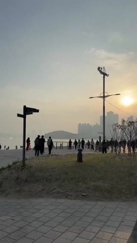 The video shows a paved walkway near a sandy area by the coast. The sun is shining brightly in the background, slightly obscured by clouds, creating a hazy atmosphere over the distant city skyline. People are strolling along the walkway, and there's a direction signpost on the left.

A woman walks past, pushing a stroller with a dog inside. The camera then pans to a wider view of the walkway, where several people are walking. One person is taking a photo toward the water, where a statue and a lighthouse are visible. The camera then pans further along the walkway, past more people and toward a breakwater structure.