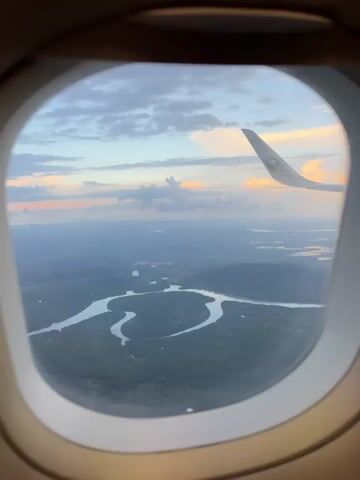 The video shows a view from an airplane window looking out at a landscape with winding rivers and lush green trees. The sky is painted with soft hues of blue, gray, and pink, suggesting it might be dawn or dusk. An airplane wing, with a logo visible, extends into the frame from the top right corner, indicating the perspective is from within the aircraft. The river system appears to meander through the forested terrain, with some bodies of water reflecting the sky's colors. The overall impression is one of serene aerial observation of a natural environment during a transitional time of day.