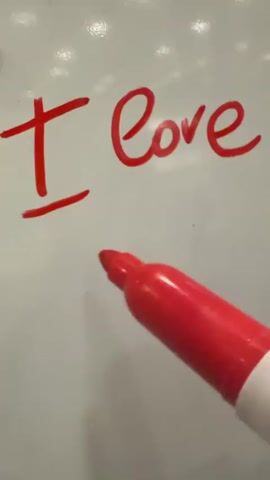 A hand holding a red marker writes "I love you" on a white board, adding a heart at the end. The words are written in cursive. The whiteboard has a glossy, slightly textured surface.