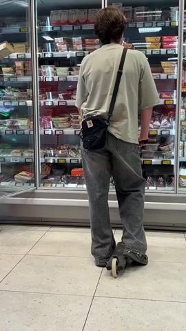 The video shows two young men in a grocery store, standing in front of a refrigerated display case filled with food products. The man on the right is facing away from the camera, wearing a light green t-shirt, grey jeans, and a black crossbody bag. He is standing on a black scooter with two wheels, one of which is partially visible. The man on the left is facing the display case, wearing a black and white jacket, grey shorts, and tan slides. He appears to be reaching into the display case. The setting is indoors, likely a supermarket, with tiled flooring and bright artificial lighting. The food products in the display case are packaged and appear to be refrigerated meals or deli items. There is no indication of the time of day or weather outside. The mood appears casual and everyday, with no discernible emotions. No visible text is present in the video.