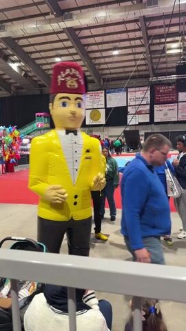 A large, inflatable doll resembling a man in a yellow tuxedo and red fez stands prominently in a crowded indoor event. The doll has pale skin and blue eyes and is adorned with a fez bearing the name "ALADDIN." People of various ages, including children, mill around the doll. Some interact with the doll, while others simply walk by. The environment appears to be an event hall or convention center, with advertisements and other displays visible in the background. A metal barrier partially obscures the foreground of the shot. The overall impression is of a festive and busy event, possibly a fair or convention.