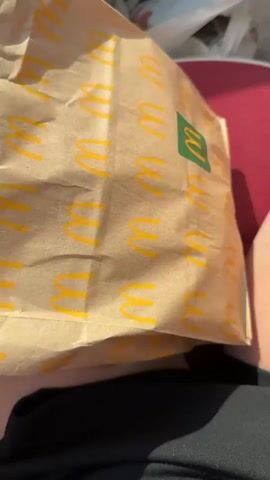 The video shows a close-up view of a McDonald's paper bag, with the iconic golden arches logo visible on it. A person's hand reaches into the bag, revealing what appears to be food items inside, possibly french fries based on the color and texture. The background is blurred, but suggests an outdoor or car setting with sunlight. The person is wearing dark clothing. The video focuses on the action of retrieving food from the bag.