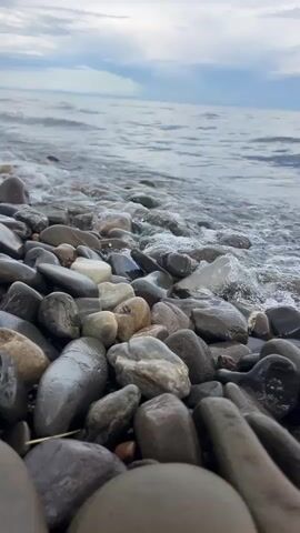 The camera is positioned at a low angle on a rocky beach, looking out over the water. The foreground is filled with smooth, wet, dark grey and brown pebbles and stones. Small waves lap against the shore, covering the rocks with water and then receding. The sky above the water is overcast, with patches of lighter clouds. The water appears calm and grey under the cloudy sky. The horizon line is visible in the distance. The sound of waves can be heard throughout the video.