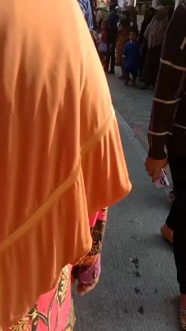 The video shows a group of people walking down a street, likely in a procession or parade. In the foreground, a person wearing an orange headscarf and a patterned skirt is visible from the side. Further down the street, a person in a blue t-shirt with white writing is walking. A child in a blue shirt is also visible. Several other people are walking in the background, some of whom are wearing traditional attire, including a person in a black and gold outfit with elaborate headwear. Flags and decorations are strung across the street. The ground appears to be paved, with some chalk markings. The overall atmosphere seems festive.