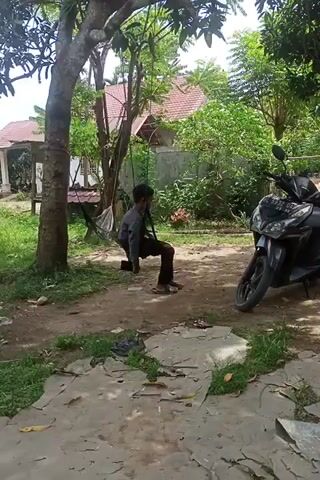 A person in a dark jacket and pants sits on a rope swing hanging from a tree. To the right of the swing is a parked black scooter. Behind the person are lush green trees and a house with a red tile roof. The ground is dirt and gravel next to a concrete path. The camera is slightly tilted.