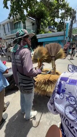 A person wearing a patterned hat, glasses, a purple long-sleeved shirt, a fanny pack, and blue jeans stands facing to the left. They are holding a hose with a blue nozzle in their hands. In front of them is a cart filled with brooms made of straw. The brooms are arranged in a circular fashion, with some upright and others lying down. Behind the person, a man in a blue shirt is riding a motorcycle. The background shows a street with buildings, trees, parked motorcycles, and other people walking. The scene appears to be outdoors on a sunny day.