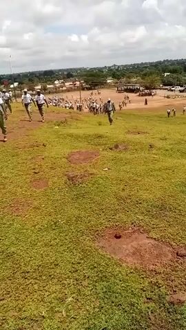 A medium-range shot shows a group of people walking down a grassy hill, towards a large group of people gathered in the distance. The people in the foreground appear to be wearing uniforms consisting of white shirts and beige pants, while the individuals in the distance seem to be similarly dressed. The background reveals a somewhat developed area with buildings, trees, and vehicles visible. The sky is partly cloudy. The ground in the foreground is green grass with patches of bare soil. The overall impression is of an outdoor gathering or event, possibly involving uniformed individuals or participants.