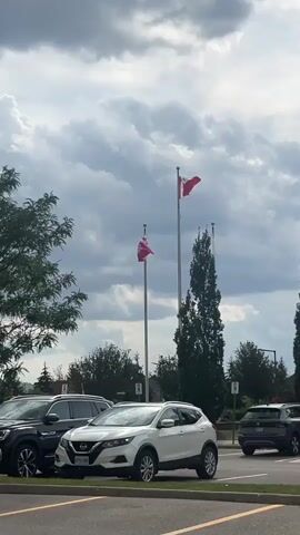 The video shows an outdoor scene with a cloudy sky. In the foreground, there is a parking lot with several cars. A white SUV is parked prominently in the center. To the left, a dark SUV is parked next to it. In the background, there are trees and flagpoles. Two flags are visible on flagpoles: the Canadian flag is flying on the taller flagpole, and another flag, possibly a provincial flag, is on a shorter flagpole to the left. The video appears to be a static shot or a very slow pan.