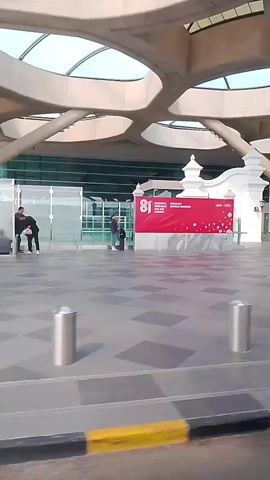 The video shows an exterior view of an airport terminal. In the foreground, a patterned walkway with a yellow and black striped border is visible. Two cylindrical trash cans are placed on the walkway. In the middle ground, a large red banner with white text and graphics is displayed, with the number "81" prominently featured. To the right of the banner, a white structure with ornate pillars stands. Beyond this, the entrance to the airport is visible, with the word "YOGYAKARTA" written above it in large letters. People are seen walking and standing in the background. The architecture of the terminal features a distinctive, curved roof structure with visible supports. The lighting suggests it is daytime.