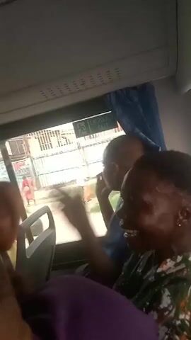 The video captures the interior of a bus from a passenger's perspective, likely in Aba, Abia, Nigeria, as indicated by the context. Several people are visible inside, with two adults prominent in the foreground looking out the window. One adult, wearing a blue shirt and with a shaved head, is actively gesturing or pointing towards something outside. The other adult, in a patterned shirt, is smiling or laughing, suggesting a lighthearted mood.

Outside the bus, an urban or semi-urban scene unfolds, with buildings and fences visible. A person dressed in red is seen walking on what appears to be a street or pathway. Two black plastic bags are also visible on the ground outside. Above the bus window, an "EMERGENCY EXIT" sign is clearly legible. The scene is illuminated by daylight, and there is singing or music playing in the background, adding to the atmosphere inside the bus.