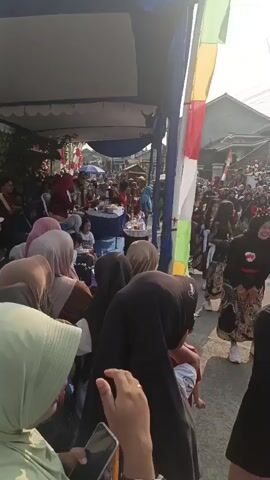 The video shows a crowded outdoor event under a large tent. People are gathered, many wearing headscarves, and some are observing a table set up in the distance with food items. Colorful flags are displayed, and the environment appears to be a street with buildings visible in the background. The sun is shining, casting shadows. The overall atmosphere suggests a community gathering or celebration.