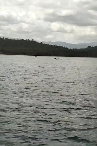 The video shows a wide shot of a body of water under a cloudy sky. In the distance, a tree-covered hillside slopes down towards the water, with a few faint mountains visible beyond. Two small boats are present on the water, one further away than the other. The water surface is rippled, indicating some movement. The overall atmosphere is calm and somewhat overcast.