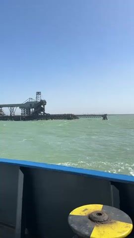 A daytime shot shows a large body of light green, wavy water with a clear, bright blue sky above. In the distance, an industrial port facility is visible on the horizon, featuring large metal structures, possibly conveyors or cranes, and what appears to be piles of material or cargo on the dock. The foreground shows the deck and railing of a boat or ship, with a dark blue hull and a light blue stripe along the top edge of the railing. Two yellow and black striped bollards (or cleats) are prominent in the lower part of the frame, indicating the vessel's presence. The water near the boat shows a slight wake, suggesting movement.