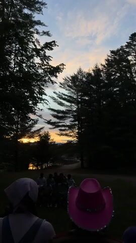 A group of young people sits in a circle around a bonfire as the sun sets behind a lake, casting a warm glow over the scene. Trees surround the clearing, and the sky is filled with hues of orange, pink, and blue. Two individuals, their backs to the camera, are in the foreground, one wearing a pink bandana and the other a bright pink cowboy hat. The video captures a moment of camaraderie and natural beauty.