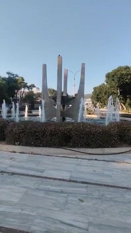 A vertical video shows a fountain in a public space. The fountain has a central abstract sculpture made of concrete and metal. Water jets spray from multiple points around the fountain's circular basin. The basin is surrounded by a thick hedge. The ground around the fountain is paved with large marble tiles. Trees and buildings are visible in the background, and the sky is clear and blue. A bird is perched on top of the central sculpture.