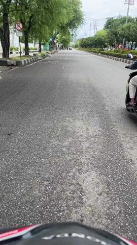 A POV shot from a motorcycle shows a person on a scooter ahead in the distance. The person is wearing a helmet and a dark jacket. The scooter is green and white. The road is paved and has some wet patches. Trees line the left side of the road. Buildings are visible in the background on the right side of the road. The sky is overcast. The motorcycle continues down the road, passing by a pedestrian crossing. The scene suggests an urban or suburban environment on a cloudy day.