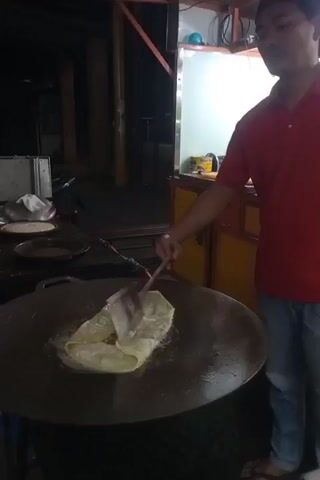 A person is shown cooking a square-shaped food item in a large, round, dark-colored pan. The food item is layered and appears to be dough with filling. It is being cooked in oil, which is sizzling around it. The person is using a long-handled metal spatula to move and flip the food item. The cooking is taking place outdoors, likely at a food stall, as indicated by the presence of a food cart or stall structure in the background. The stall has a yellow and orange color scheme with shelving and a white countertop. The lighting suggests it is nighttime or early morning. There are other items on the countertop and in the background, including what appear to be packaged goods. The overall environment appears to be a street or market setting.