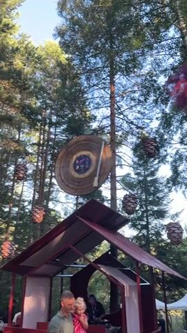 A wide shot shows a forest setting with a DJ booth and people. In the foreground, a man holds a young child. Behind them is a red DJ booth with two DJs playing music. Above the DJ booth, a large record player is hanging from a tree. The area is decorated with pinecone-shaped ornaments. The overall atmosphere suggests an outdoor event or festival.