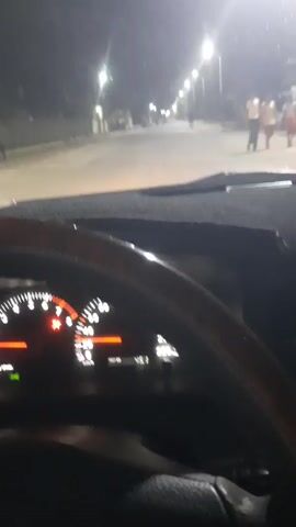 A first-person perspective video shows a person driving a car at night. The car's dashboard is visible, with a speedometer reading approximately 45 km/h and a tachometer reading around 1000 RPM. The headlights are on, illuminating a street lined with streetlights. Several people and a motorcycle are visible in the distance on the road. The environment appears to be a town or village.