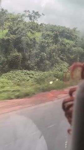 The video captures a view from inside a vehicle, looking out a side window. The landscape is characterized by lush green vegetation covering rolling hills. A dirt road, appearing somewhat uneven and red-hued, runs alongside the vehicle. In the foreground, a portion of the vehicle's interior is visible, including what appears to be a person's hair. The sky is overcast with clouds. The overall impression is of a journey through a natural, possibly rural or undeveloped, environment.