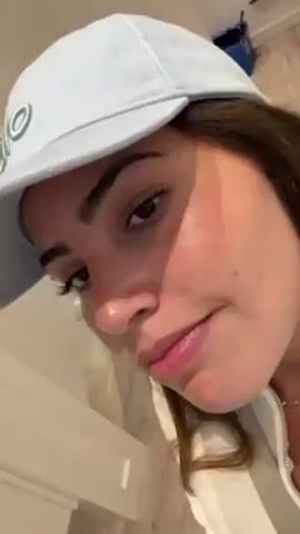 A close-up shot shows a light-skinned woman wearing a light blue baseball cap with a small design on the front. She has brown hair and brown eyes. She is wearing a white jacket with a zipper. Her lips are slightly puckered, and she appears to be looking at the camera. A blue toolbox is visible in the background.  She moves her hand to adjust her hat.