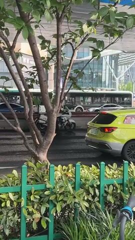 The video depicts a wet street scene in an urban setting, likely in China given the presence of Chinese characters. Initially, a police officer on a white motorcycle is visible, with the word "police" written in Chinese on the vehicle. A green car is nearby, and several buses pass in the background. The camera then pans upwards, revealing an overpass, and a person wearing a hoody is seen standing near the edge of the road. The environment includes commercial buildings and billboards in the distance. The road appears to be wet from rain.