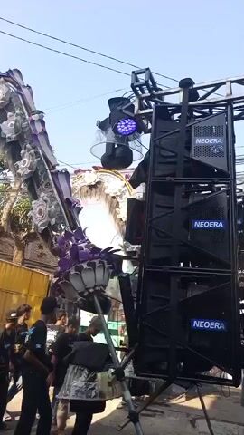 The video shows a large, elaborate parade float that appears to be decorated with large, ornate floral or star-shaped ornaments and dark, feathery accents. The float is also equipped with multiple speakers, stage lighting, and what looks like a spotlight or moving head light. Several people are visible around the float, some appearing to be associated with its operation or decoration, while others seem to be onlookers. The environment suggests an outdoor setting, possibly a street, with buildings and power lines in the background. The overall scene appears to be a public event or celebration.
