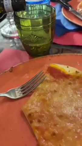 Here is a description of the video:

The video shows someone pouring a bottle of what appears to be olive oil into a green glass. There is a slice of pizza on an orange plate in the foreground. A fork is also visible on the plate. Other plates, napkins, and utensils are present on the table, suggesting a dining setting.