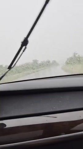 The video shows a view from inside a car on a rainy day. The windshield wipers are active, clearing the view of the road ahead. The road appears wet and there is reduced visibility due to the rain and possibly fog. Trees and vegetation line the sides of the road. The dashboard of the car is visible in the foreground. The overall impression is of driving in inclement weather.