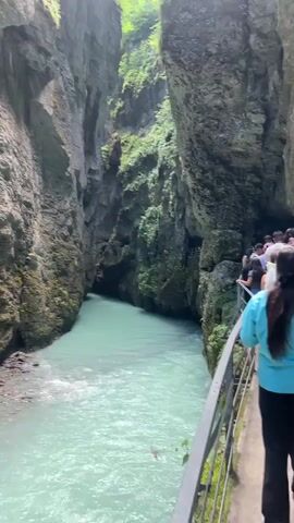 A person is filming from a walkway next to a bright turquoise river flowing through a narrow canyon. The walkway has a metal railing. On the walkway, a group of people are walking away from the camera. The canyon walls are steep and rocky, with some vegetation growing on them. The river is wide and appears to be moving quickly. The camera briefly pans to the right, showing more of the canyon wall.