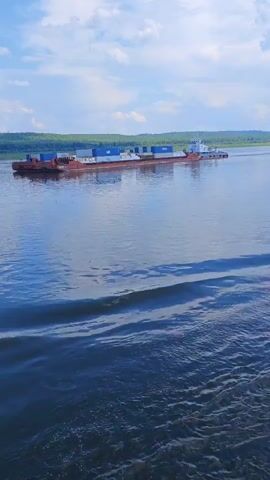 The video shows a wide, calm body of water with a barge carrying shipping containers moving from left to right across the frame. The sky above is partly cloudy. The water in the foreground has ripples and wakes, indicating movement. The barge is long and flat, with a tugboat attached to its rear, pushing it. The containers on the barge are primarily blue and white. The shoreline in the distance is lined with trees. The perspective is from a lower vantage point, likely from another moving vessel.