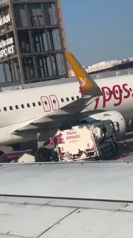 An airplane is parked on the tarmac. A white truck with red accents is parked next to the airplane's wing. The truck has "Pegasus Air" written on its side. Two people are visible near the truck and the airplane. In the background, a control tower with "Dalaman Airport" written on it is visible. The sky is clear and the sun is shining.