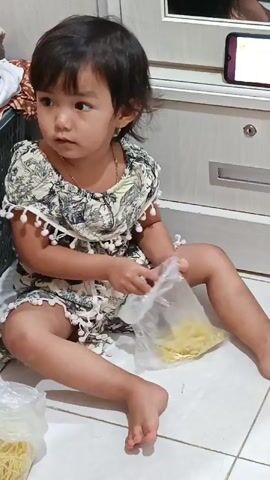 A young child with dark hair, wearing a patterned dress with pom-pom trim, sits on a tiled floor. The child is holding a plastic bag containing yellow noodles and appears to be playing with them using a fork. Another plastic bag of noodles is visible on the floor to the left of the child. In the background, a white cabinet with a lock is partially visible, and a smartphone is propped up on top of it, displaying an image. The child is looking down at the noodles and the bag. The setting appears to be indoors.