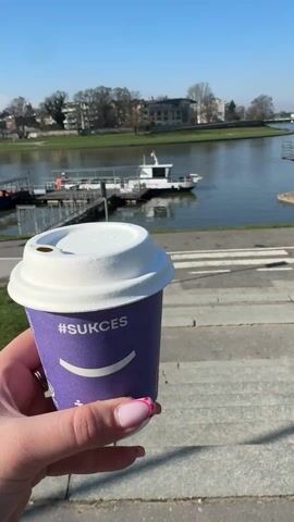 The video starts with a close-up of a hand holding a purple coffee cup with the hashtag "#SUKCES" and a smiley face on it. The background is a view of a river, a pedestrian crossing, and some buildings. The camera then pans to show the river with a boat docked at a pier. The camera pans further to the left, revealing a paved walkway, grass, and more boats along the riverbank. A person is walking on the path. The sky is clear and blue.